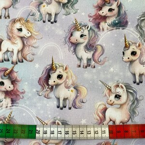 May include: A purple fabric with a repeating pattern of white unicorns with rainbow manes and tails. The unicorns have gold stars on their bodies and are surrounded by white stars and rainbow arcs.
