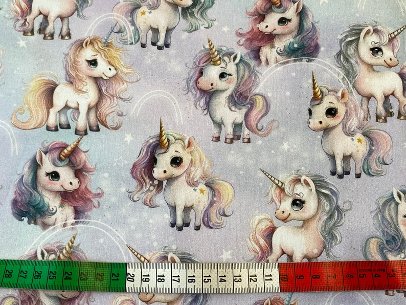 May include: A purple fabric with a repeating pattern of white unicorns with rainbow manes and tails. The unicorns have gold stars on their bodies and are surrounded by white stars and rainbow arcs.