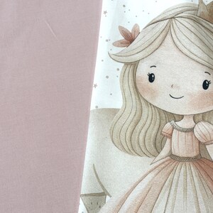 Puede incluir: Un panel de tela con una ilustraci&oacute;n impresa de un personaje con cabello rubio largo, una peque&ntilde;a corona y un vestido color melocot&oacute;n. El personaje est&aacute; rodeado de peque&ntilde;as estrellas. La tela se combina con un textil rosa empolvado.