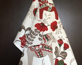 Puede incluir: Un paño de cocina blanco con un diseño de caballo Dala escandinavo rojo y gris. El caballo está rodeado de flores rojas y verdes.