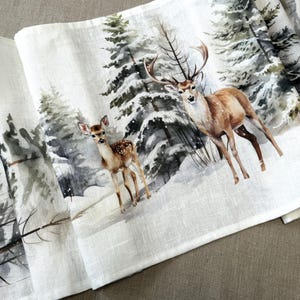May include: White linen table runner depicting a winter forest scene with deer. The design includes a buck with large antlers and a fawn standing in a snowy landscape with evergreen trees. The runner is folded.