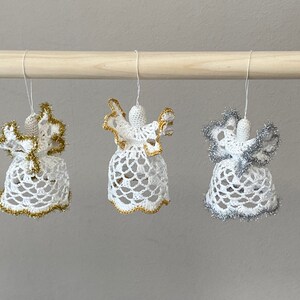 May include: Three crocheted angel ornaments hanging from a wooden dowel. The angels are white with gold and silver trim.