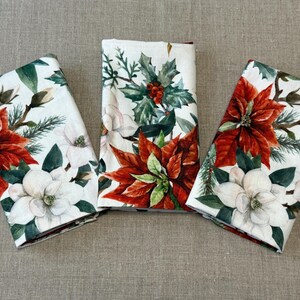 Puede incluir: Tres servilletas de lino blanco con un dise&ntilde;o floral con poinsettias rojas, flores de magnolia blancas y hojas de acebo verdes.