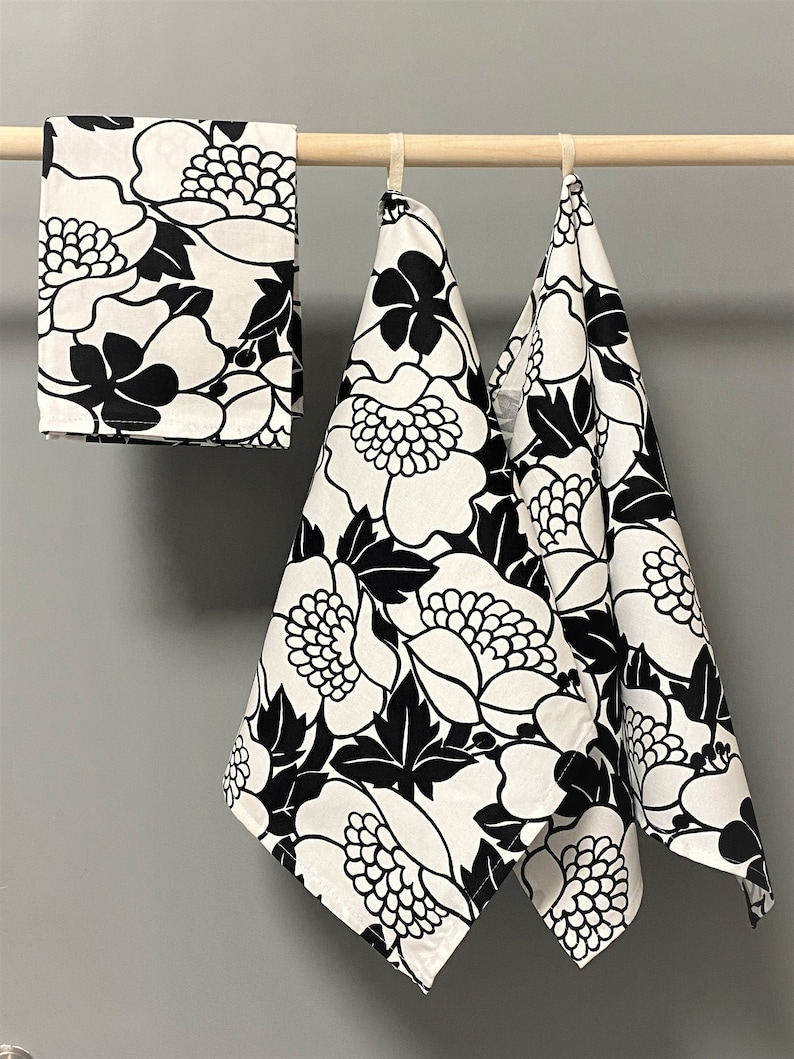 May include: Three white kitchen towels with a black floral pattern. The towels are hanging on a wooden rod.