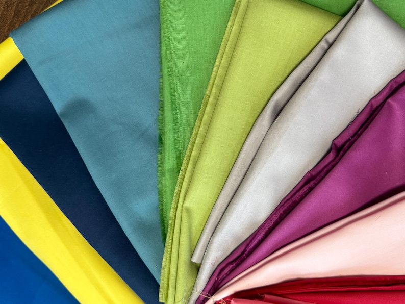 May include: A fan of colorful fabric swatches in shades of yellow, blue, teal, green, silver, purple, and pink. The fabric appears to be a smooth, possibly silk or satin material, with a slight sheen. The arrangement showcases a variety of colors.