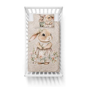 May include: A crib set featuring a watercolor illustration of bunnies. The set includes a fitted sheet, a pillowcase, and a quilt. The quilt and pillowcase have a repeating pattern of small flowers. The main bunny wears a floral dress.