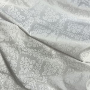 May include: A close-up of a white fabric with a floral damask pattern. The fabric is draped, showing folds and shadows. The pattern features large, stylized flowers and leaves, creating a textured appearance. The fabric has a subtle sheen.