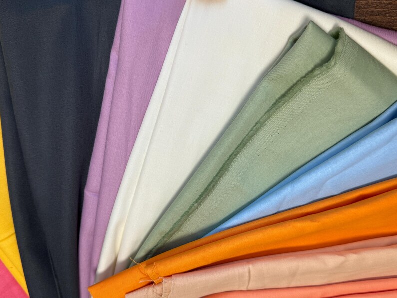 May include: A collection of folded fabric in various colors, including black, lavender, white, green, light blue, orange, and peach. The fabrics are arranged in a fan-like pattern, showcasing the different hues and textures.