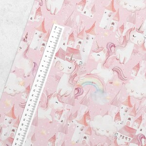 May include: Pink fabric featuring a repeating pattern of white unicorns with pink manes and wings, castles, rainbows, and clouds. A ruler is placed diagonally across the fabric, measuring up to 24 cm.