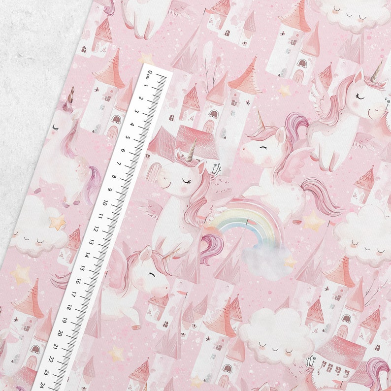 May include: Pink fabric featuring a repeating pattern of white unicorns with pink manes and wings, castles, rainbows, and clouds. A ruler is placed diagonally across the fabric, measuring up to 24 cm.