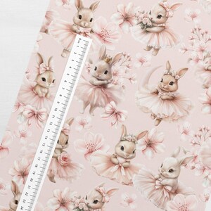 May include: A light pink fabric featuring a repeating pattern of ballet-dancing bunnies in pink tutus, bows, and floral accents. A ruler is placed on the fabric, with measurements up to 24 cm.