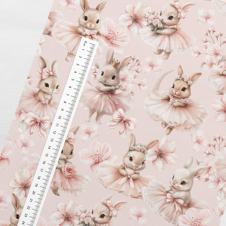 May include: A light pink fabric featuring a repeating pattern of ballet-dancing bunnies in pink tutus, bows, and floral accents. A ruler is placed on the fabric, with measurements up to 24 cm.