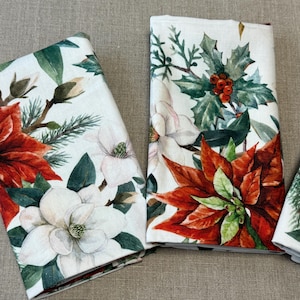 Puede incluir: Tres servilletas de lino blanco con un estampado floral con poinsettias rojas, flores de magnolia blancas y hojas de acebo verdes.