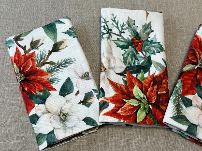 Puede incluir: Tres servilletas de lino blanco con un estampado floral con poinsettias rojas, flores de magnolia blancas y hojas de acebo verdes.