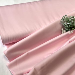 Puede incluir: Un gran rollo de tela rosa claro y suave con elegantes pliegues. Un pequeño ramo de flores blancas con hojas verdes descansa sobre la tela, añadiendo un toque de belleza natural. La tela parece suave y lujosa.