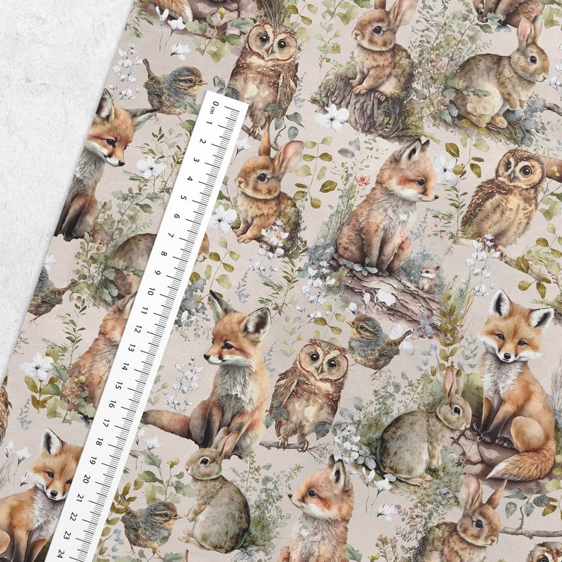 May include: A fabric with a repeating pattern of woodland animals including foxes, owls, rabbits, and birds. The animals are in shades of brown, tan, and gray, set against a light beige background with floral and foliage accents. A ruler is placed on the fabric, measuring up to 24 inches.