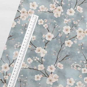 May include: A blue fabric with a white floral pattern. The fabric features a repeating pattern of delicate white flowers with pink centers, resembling cherry blossoms, on a light blue background. A white ruler is placed on the fabric, with the numbers 0 to 24 visible.