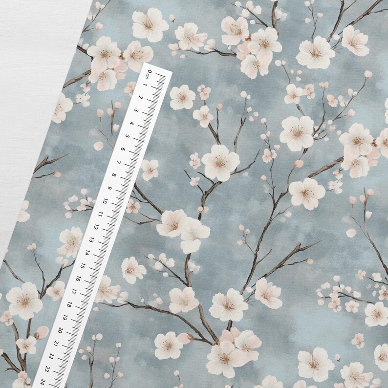 May include: A blue fabric with a white floral pattern. The fabric features a repeating pattern of delicate white flowers with pink centers, resembling cherry blossoms, on a light blue background. A white ruler is placed on the fabric, with the numbers 0 to 24 visible.