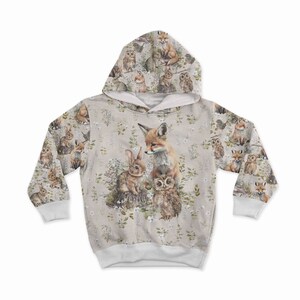 May include: A beige hooded sweatshirt featuring a woodland animal print. The design includes a fox, rabbit, and owl, surrounded by floral and foliage elements. The sleeves and hood are covered in a repeating pattern of forest creatures.