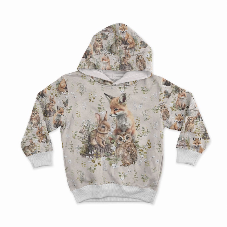 May include: A beige hooded sweatshirt featuring a woodland animal print. The design includes a fox, rabbit, and owl, surrounded by floral and foliage elements. The sleeves and hood are covered in a repeating pattern of forest creatures.