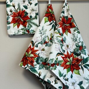May include: Three white kitchen towels with a floral pattern featuring red poinsettias, white magnolia flowers, and green holly leaves.