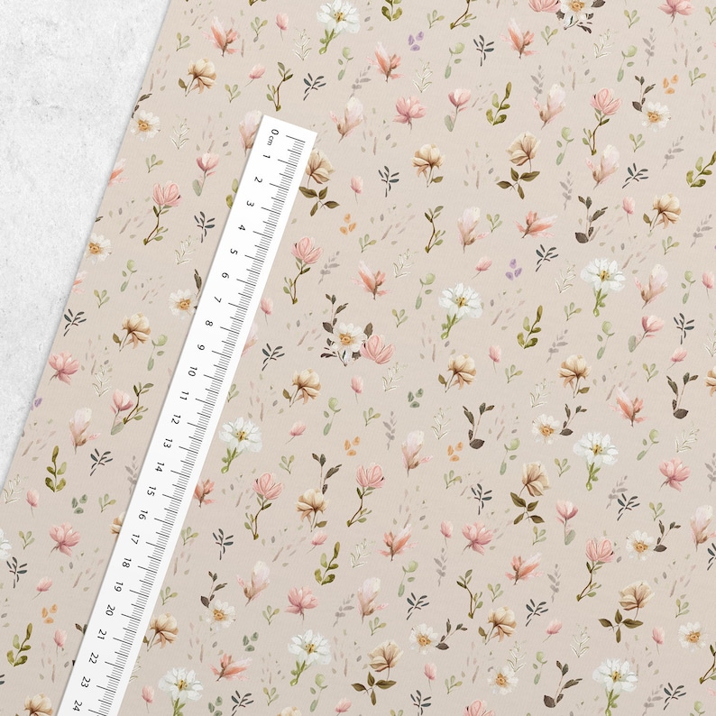 May include: A beige fabric with a repeating pattern of small pink and white flowers, green leaves, and brown dots. The fabric is measured with a ruler in inches.