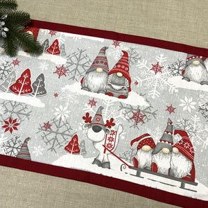 以下が含まれることがあります： 赤と白のノーム、雪の結晶、木々が描かれた、グレーの背景のクリスマスをテーマにしたテーブルランナー。 ランナーは濃い赤色の縁取りがあり、お祝いの休日の画像で飾られています。
