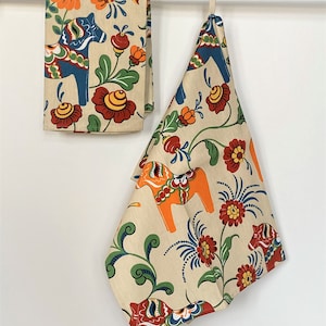Puede incluir: Dos toallas de cocina beige con un patrón floral con caballos Dala suecos coloridos. Los caballos están pintados en azul, naranja, rojo y amarillo. Las flores son rojas, naranjas, amarillas y azules.