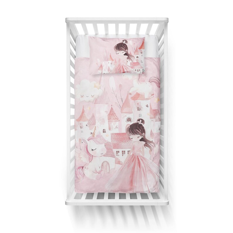 K&ouml;nnte beinhalten: Ein rosa-wei&szlig;es Babybett-Bettw&auml;sche-Set mit einem Einhorn- und Prinzessinnen-Motiv. Die Bettw&auml;sche zeigt ein rosa Schloss, Wolken und Sterne. Der Kissenbezug zeigt eine Prinzessin mit braunen Haaren und einem rosa Kleid.