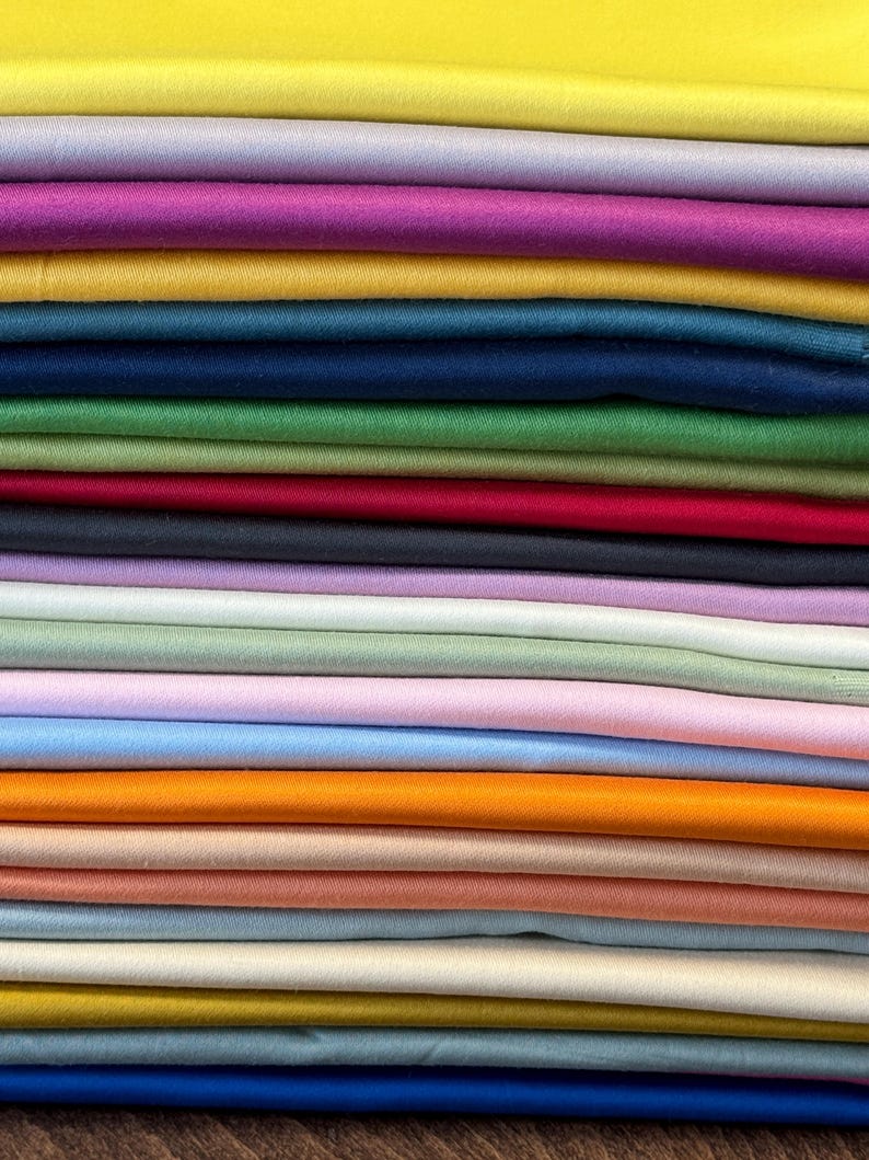 May include: A stack of folded fabric in various colors, including yellow, purple, pink, teal, green, red, and blue. The fabric appears to be a smooth, solid material, possibly cotton or silk, suitable for sewing or crafting projects.