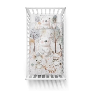 May include: A white crib bedding set with a woodland theme. The bedding features a white background with brown trees, a white bear, a rabbit, a mouse, and ducks. The bedding is made of soft, comfortable fabric.