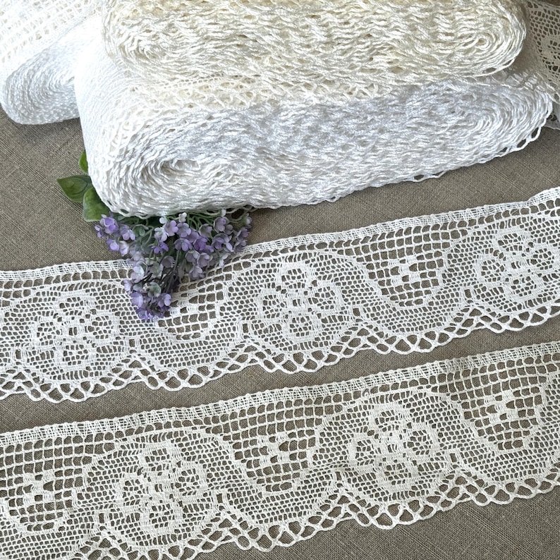 K&ouml;nnte beinhalten: Nahaufnahme von wei&szlig;en Spitzenbortenrollen und zwei Spitzenstreifen. Die Spitzenborte hat ein florales Design. Die Spitzenrollen sind in verschiedenen Wei&szlig;t&ouml;nen gehalten. Ein Zweig lila Blumen ist in der N&auml;he der Spitze platziert.