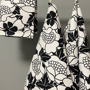 May include: Three black and white floral kitchen towels hanging on a wooden rod. The towels have a large floral pattern with black flowers and leaves on a white background.