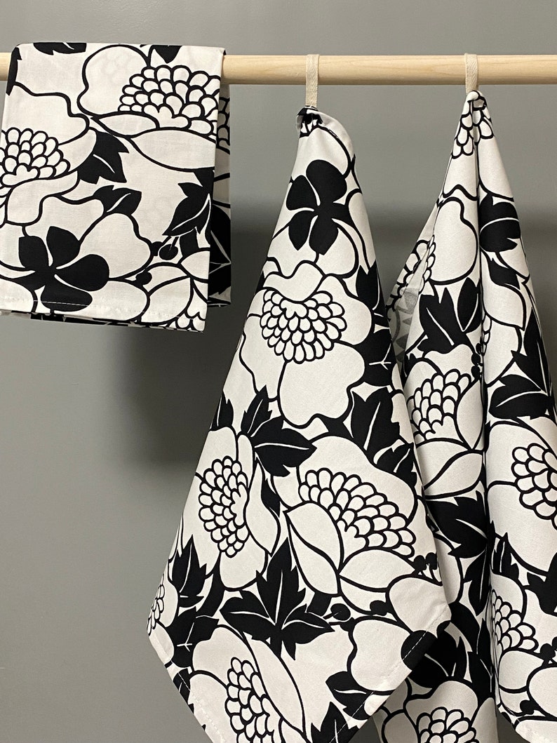 May include: Three black and white floral kitchen towels hanging on a wooden rod. The towels have a large floral pattern with black flowers and leaves on a white background.