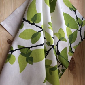 Puede incluir: Toalla de cocina blanca con un patrón de hojas verdes. Las hojas son estilizadas y tienen un contorno verde oscuro. La toalla está doblada por la mitad y colgada en una superficie de madera.