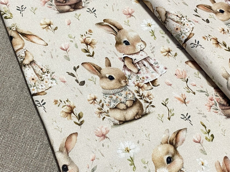 May include: A beige fabric with a repeating pattern of brown bunnies wearing clothes and flowers. The bunnies are in various poses, some are sitting, some are standing, and some are looking to the side. The flowers are in shades of pink, white, and brown.