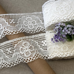 K&ouml;nnte beinhalten: Cremefarbenes H&auml;kelspitzenband mit floralem Design. Das Band ist aufgerollt und teilweise abgerollt, wodurch das komplizierte Lochmuster sichtbar wird. Ein kleiner Strau&szlig; lila Blumen liegt auf dem Band.