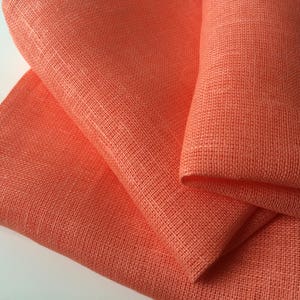 May include: A stack of three coral orange linen napkins. The napkins are made of a woven fabric with a slightly rough texture.