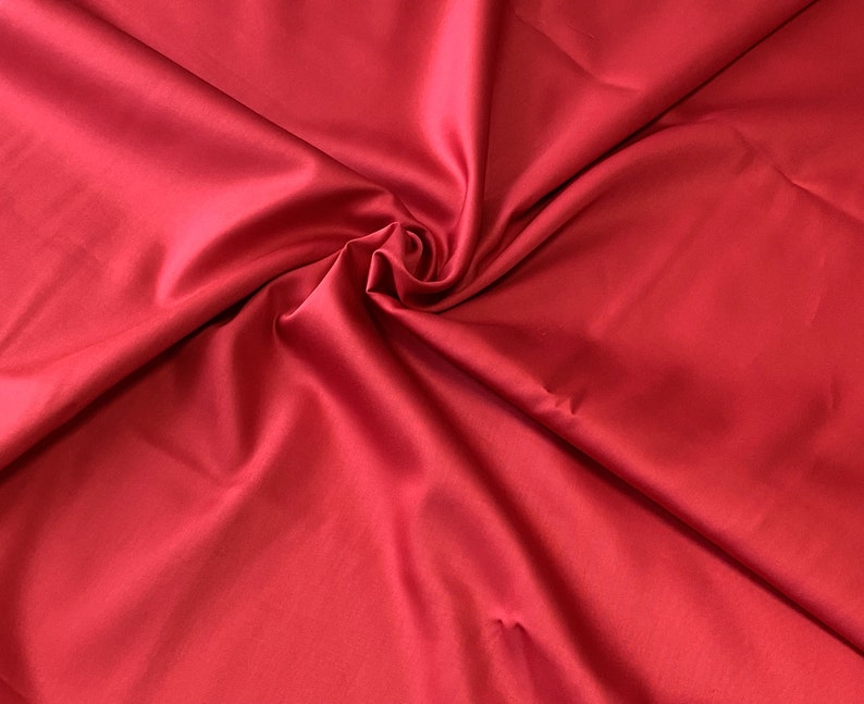 May include: A close-up shot of a vibrant red fabric with a smooth, satin-like texture. The fabric is gathered and swirled, creating soft folds and shadows. The color is a rich, solid red, with a slight sheen.