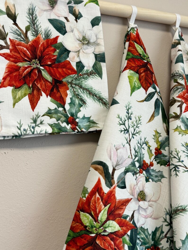 Puede incluir: Tres toallas de cocina blancas con un patr&oacute;n floral que presenta flores rojas de poinsettia, flores blancas de magnolia y bayas verdes de acebo.