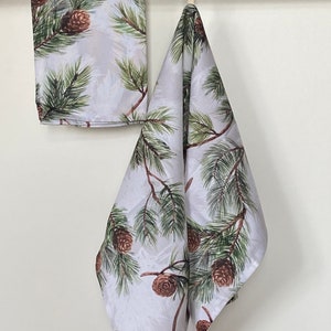 Puede incluir: Dos toallas de cocina blancas con un patrón de pino y piña. Las toallas están colgadas en una barra plateada.