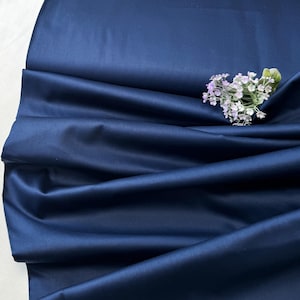 May include: A close-up shot of navy blue fabric, showing soft folds and a slight sheen. A small arrangement of delicate, pale purple flowers with green leaves sits on the fabric. The material appears to be smooth, possibly satin or silk.
