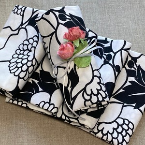 May include: Four white napkins with a black floral pattern are folded and stacked. A white ribbon with two pink flowers is tied around the top napkin.