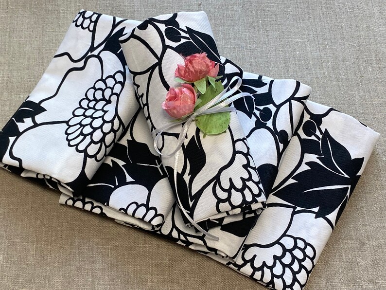 May include: Four white napkins with a black floral pattern are folded and stacked. A white ribbon with two pink flowers is tied around the top napkin.