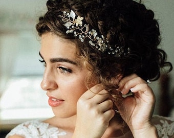 May include: A bride with curly hair wears a white lace wedding dress and a gold leaf and crystal hair crown. She is putting on a gold earring.