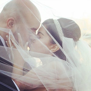 May include: A bride and groom kiss under a white veil. The bride is wearing a white dress and a tiara. The groom is wearing a black suit.