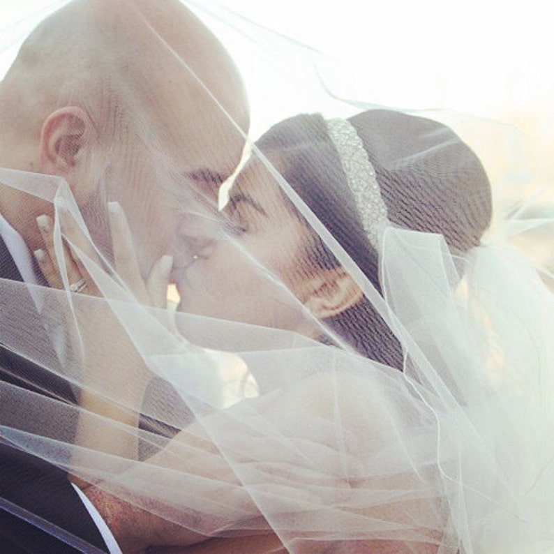 May include: A bride and groom kiss under a white veil. The bride is wearing a white dress and a tiara. The groom is wearing a black suit.
