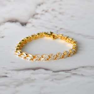 K&ouml;nnte beinhalten: Ein goldenes Armband mit einem zarten Design aus klaren, marquisef&ouml;rmigen Steinen, die in einer Reihe angeordnet sind. Das Armband liegt auf einer wei&szlig;en Marmorfl&auml;che.