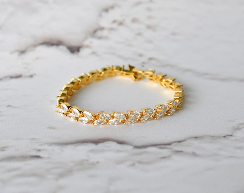 K&ouml;nnte beinhalten: Ein goldenes Armband mit einem zarten Design aus klaren, marquisef&ouml;rmigen Steinen, die in einer Reihe angeordnet sind. Das Armband liegt auf einer wei&szlig;en Marmorfl&auml;che.