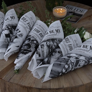 May include: Five white paper cones with black text and images. The text on the cones reads "Will you be my bridesmaid?" and "The Wedding Times". The cones are on a wooden table with a lit candle and a magazine.